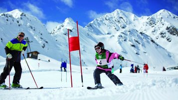 Ein Kind fährt auf Ski einen Hang im Rahmen eines Skirennens für Kinder hinunter. Daneben steht Skilegende Gustav Thöni vom Hotel Bella Vista in Südtirol.