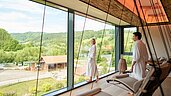 Ein Mann und eine Frau im Bademantel im Ruhebereich des Wellnessbereich mit Blick auf die Aussenanlage des Familienhotels Schreinerhof im Bayerischen Wald.