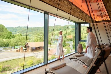 Ein Mann und eine Frau im Bademantel im Ruhebereich des Wellnessbereich mit Blick auf die Aussenanlage des Familienhotels Schreinerhof im Bayerischen Wald.