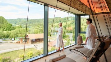 Ein Mann und eine Frau im Bademantel im Ruhebereich des Wellnessbereich mit Blick auf die Aussenanlage des Familienhotels Schreinerhof im Bayerischen Wald.