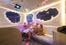 Familie sitzt lachend im farbenfroh beleuchteten Dampfbad des Zauchenseehofs, umgeben von spielerischen Wolken- und Tierprojektionen an den Wänden.