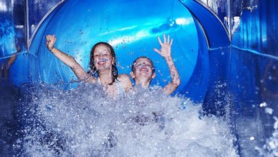 Im Familienhotel Schwarzwald - zwei lachende Kinder rutschen in rasantem Tempo eine blaue Wasserrutsche hinunter und landen mit spritzendem Wasser im Becken.