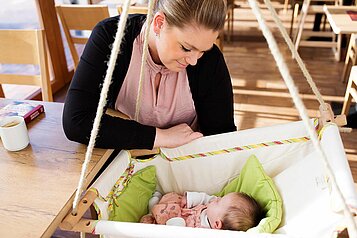 Baby liegt in einer Babyschaukel im Restaurant des Familienhotels Landhuus Laurenz und die Mutter sitzt daneben am Tisch und umsorgt ihr Kind.