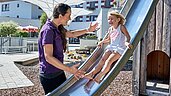 Kind rutscht eine Rutsche auf dem Outdoor Spielplatz des Familienhotels Amiamo hinunter. Eine freundliche Kinderbetreuerin hilft ihr dabei.