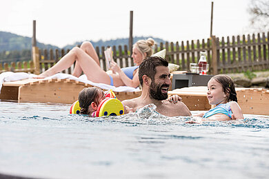 Vater mit seinen zwei Kindern im Aussen-Pool eines Hotels. Mutter liegt gemütlich auf einer Liege und liest ein Buch 