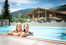 Zwei Mädchen lehnen am Beckenrand eines Pools, im Hintergrund ein Holzhaus und grüne Berge, im Familienhotel Spa- & Familien-Resort Krone im Allgäu.