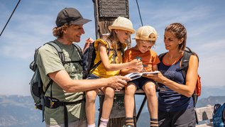 Familie mit zwei Kindern am Gipfel eines Berges in Tirol mit einem Gipfelbuch in der Hand.