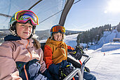 Zwei Teens sitzen im Winter im Sessellift und fahren die Skipiste hinauf.