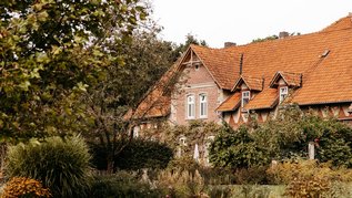 Rotes Backsteinhaus mit orangefarbenem Ziegeldach, umgeben von dichtem Garten mit blühenden Stauden und Büschen.