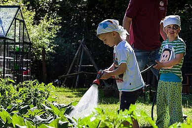 Zwei als Piraten verkleidete Kinder stehen im Gemüsegarten des Familienhotels Borchard's Rookhus an der Mecklenburgischen Seenplatte und gießen mit einem Gartenschlauch die Pflanzen.