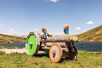 Ein Kind sitzt auf einem Holztraktor, im Hintergrund sieht man einen kleinen Bergsee.