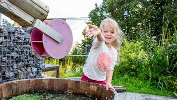 Ein fröhliches Mädchen spielt an einem Wasserspielplatz nahe des Familienhotels Engel mit hölzerner Wasserrinne und Bottich