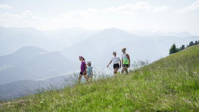 Familie wandert über eine grüne Bergwiese mit weitem Blick auf die Alpen beim Familienhotel Sonne Bezau.