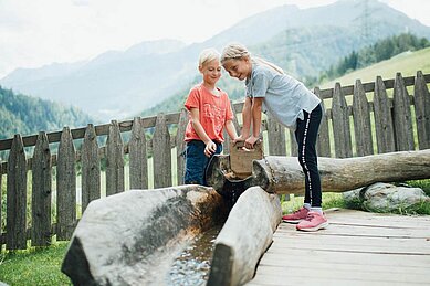 Ein Junge und ein Mädchen spielen auf der Abenteuerspielwiese des Kinderhotels Sailer mit Wasser.