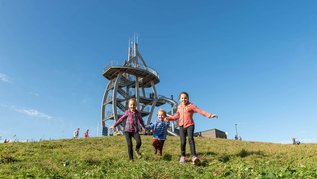 Wandern in der Rhön: Kinder laufen über eine Wiese im Hintergrund der Aussichtsturm auf dem Naturlehrpfad "Ellenbogen"