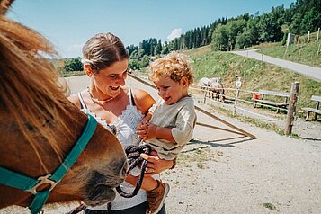 Mutter mit ihrem Kind auf dem Arm beim Streicheln der Pferde im Familien Resort Petschnighof