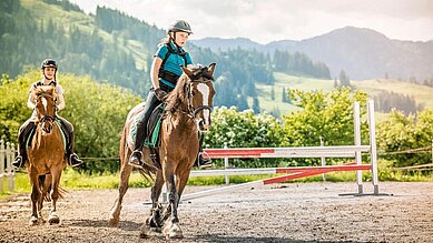 Zwei Mädchen sitzen auf Pferden und reiten auf dem Reitplatz des Familienhotels Landgut Furtherwirt.