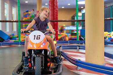 Ein fröhliches Mädchen fährt ein elektrisches Kinder-Motorrad auf einer Indoor-Rennbahn im Familienhotel Schreinerhof im Bayrischen Wald, mit einem bunten Spielbereich im Hintergrund.