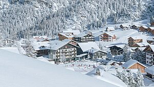 Atemberaubende Winteransicht eines Familienhotels inmitten einer verschneiten Landschaft mit dicht verschneiten Bäumen und einer Skipiste im Vordergrund, die ein idyllisches und einladendes Urlaubsziel für Familien darstellt.