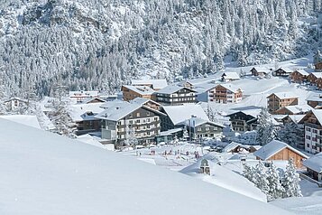 Atemberaubende Winteransicht eines Familienhotels inmitten einer verschneiten Landschaft mit dicht verschneiten Bäumen und einer Skipiste im Vordergrund, die ein idyllisches und einladendes Urlaubsziel für Familien darstellt.